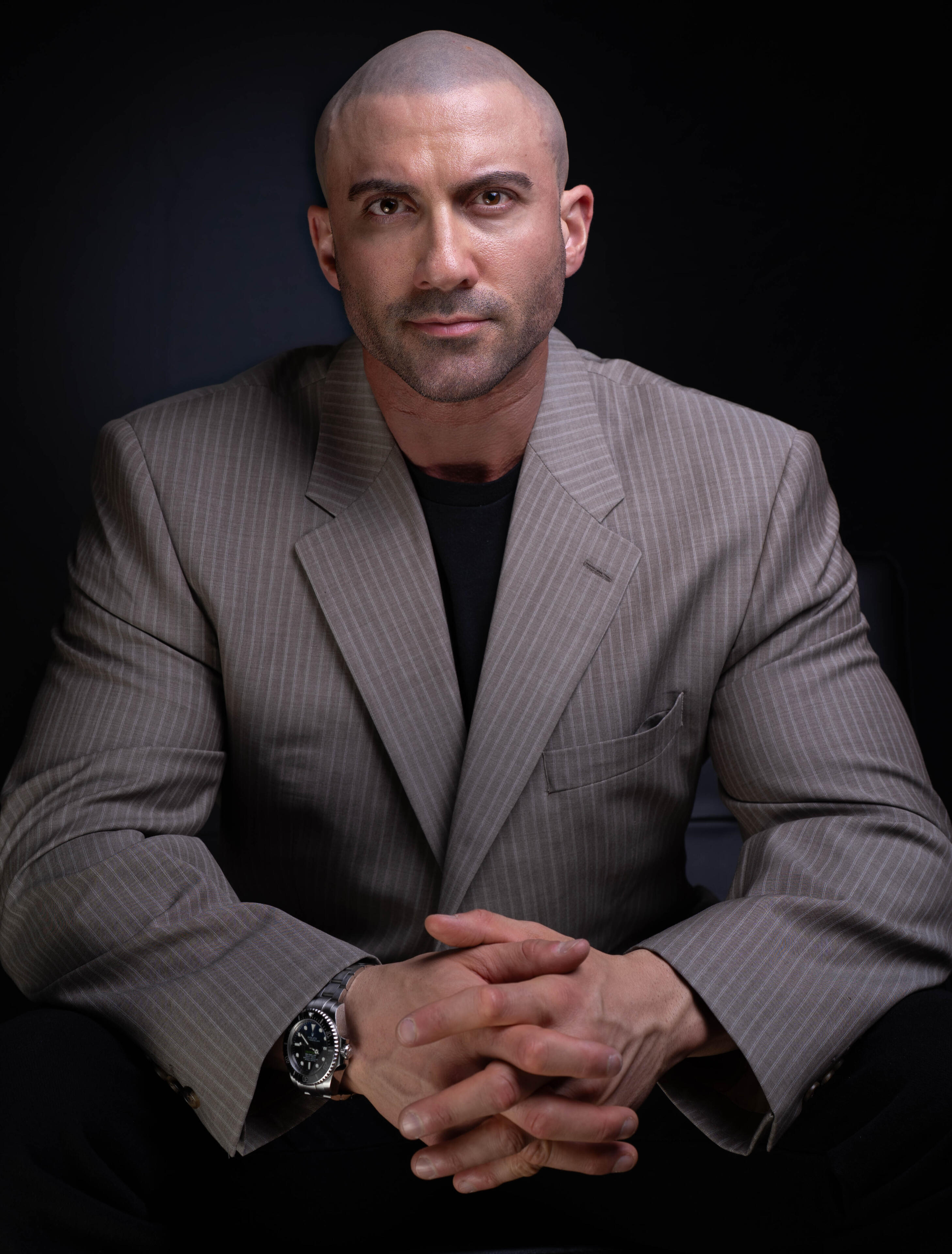 Thomas (Tom) Rooney Thomas Rooney (Tom Rooney), Author, Founder & CEO, and Former Headliner Performer, sitting with black background wearing grey business suit with hands clasped looking at camera.